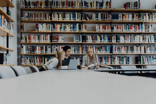Drei Studierende lernen gemeinsam in der Bibliothek vor großem Büchrregal
