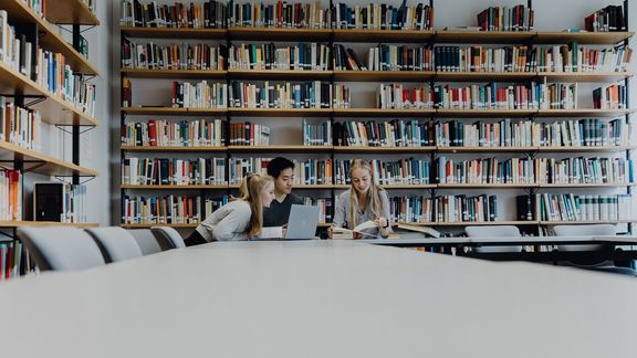 Drei Studierende lernen gemeinsam in der Bibliothek vor großem Büchrregal
