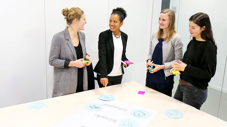 Eine Gruppe von vier jungen Frauen steht um einen Tisch, auf dem ein Flipchart Papier liegt
