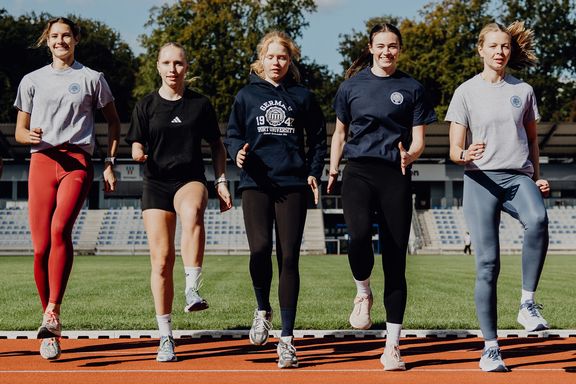 Foto von Sportlerinnen die auf der Laufbahn nebeneinander stehen und Aufwärmübungen machen
