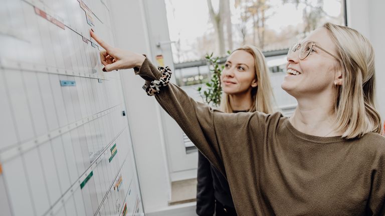 Foto von zwei Frauen die einen Terminkalender auf einem Whiteboard betrachten
