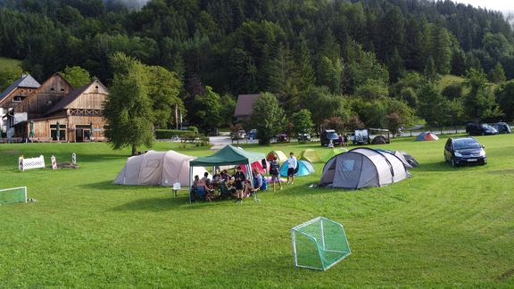 Mehrere Zelte stehen auf einer großen grünen Wiese vor einem bewaldeten Hügel. Unter einem Pavillon sitzt eine Gruppe von Menschen zusammen. Im Hintergrund sind Holzhäuser, parkende Autos und weitere Zelte zu sehen. Auf der Wiese stehen außerdem kleine Fußballtore.
