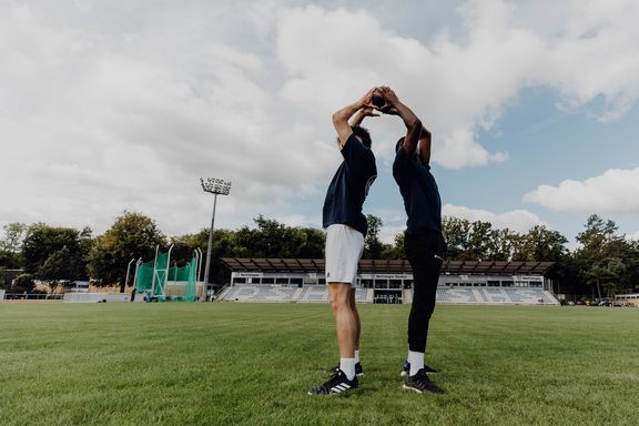 Photo of two men passing a ball to each other head-to-head, back-to-back, on a grass field
