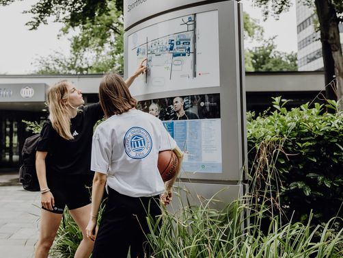 Zwei Studierende zeigen auf den Wegweiser im Eingangsbereich der Deutschen Sporthochschule
