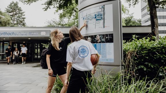 Zwei Studierende zeigen auf den Wegweiser im Eingangsbereich der Deutschen Sporthochschule
