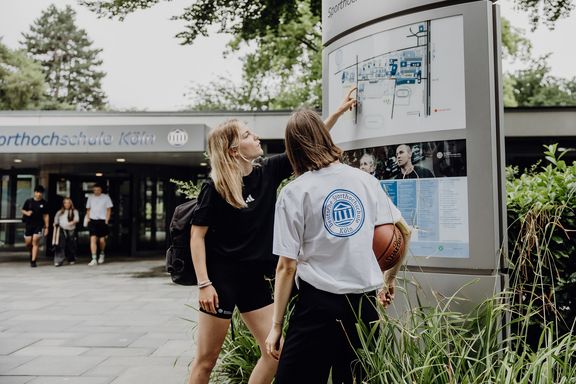 Zwei Studierende zeigen auf den Wegweiser im Eingangsbereich der Deutschen Sporthochschule
