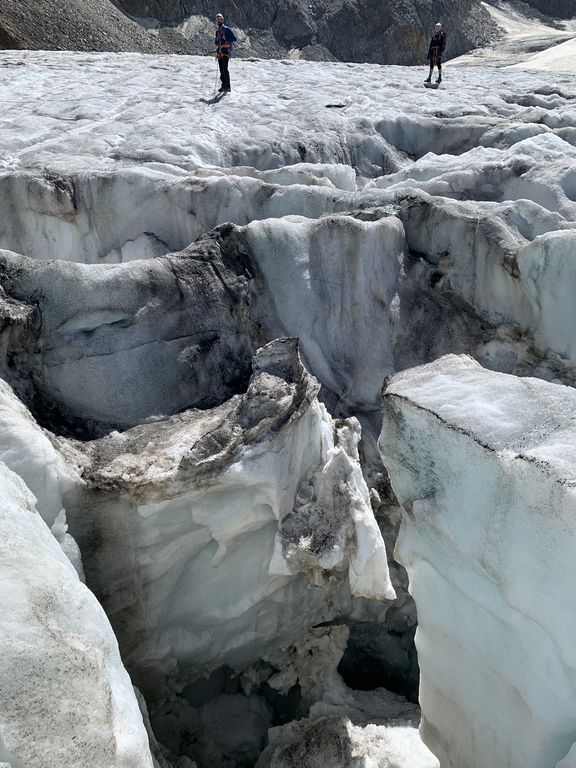 Zwei Personen stehen auf einem zerklüfteten Gletscher mit tiefen Spalten und Eisformationen
