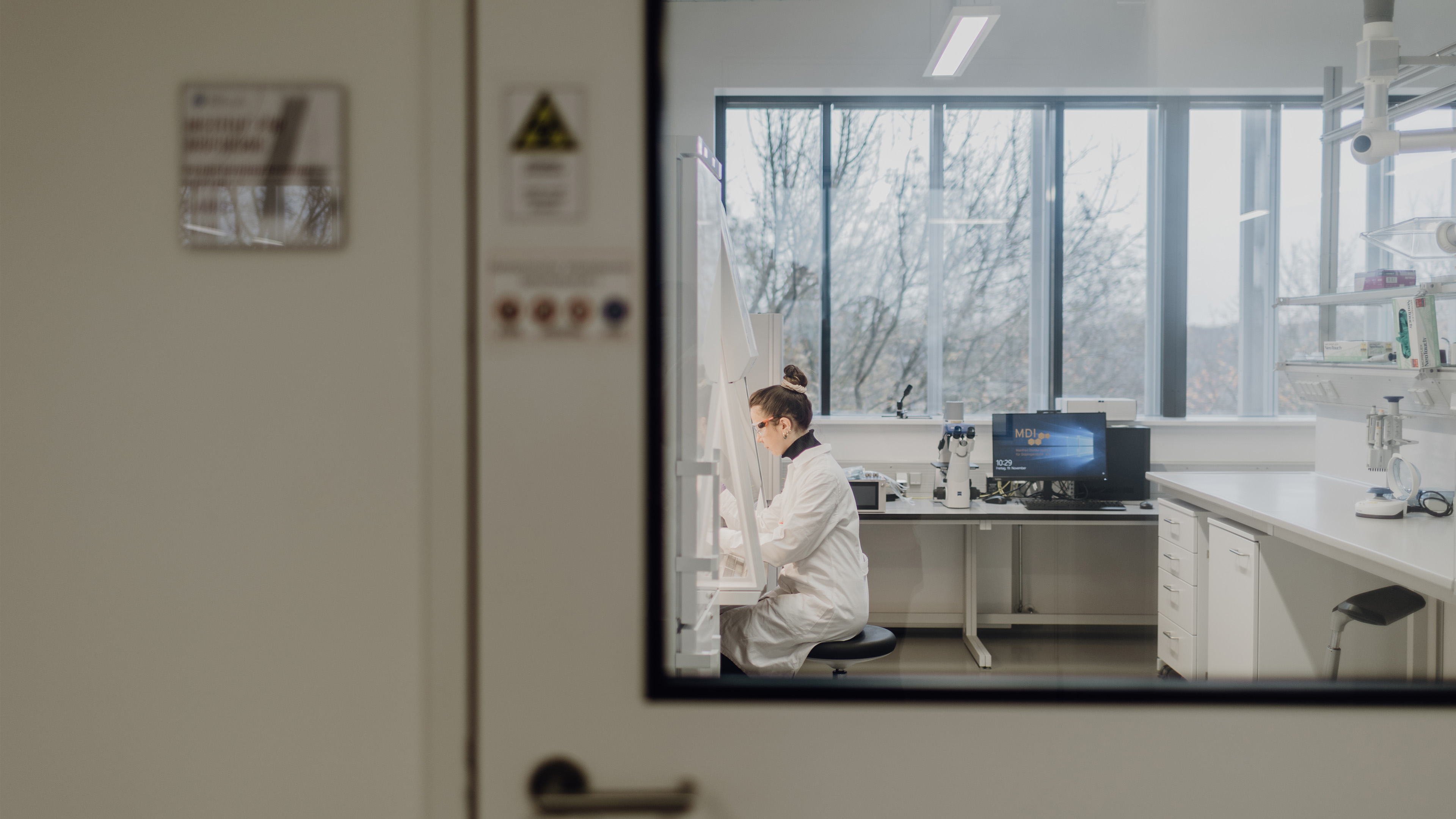 Foto durch verglaste Tür in ein Labor in dem eine Person sitzt
