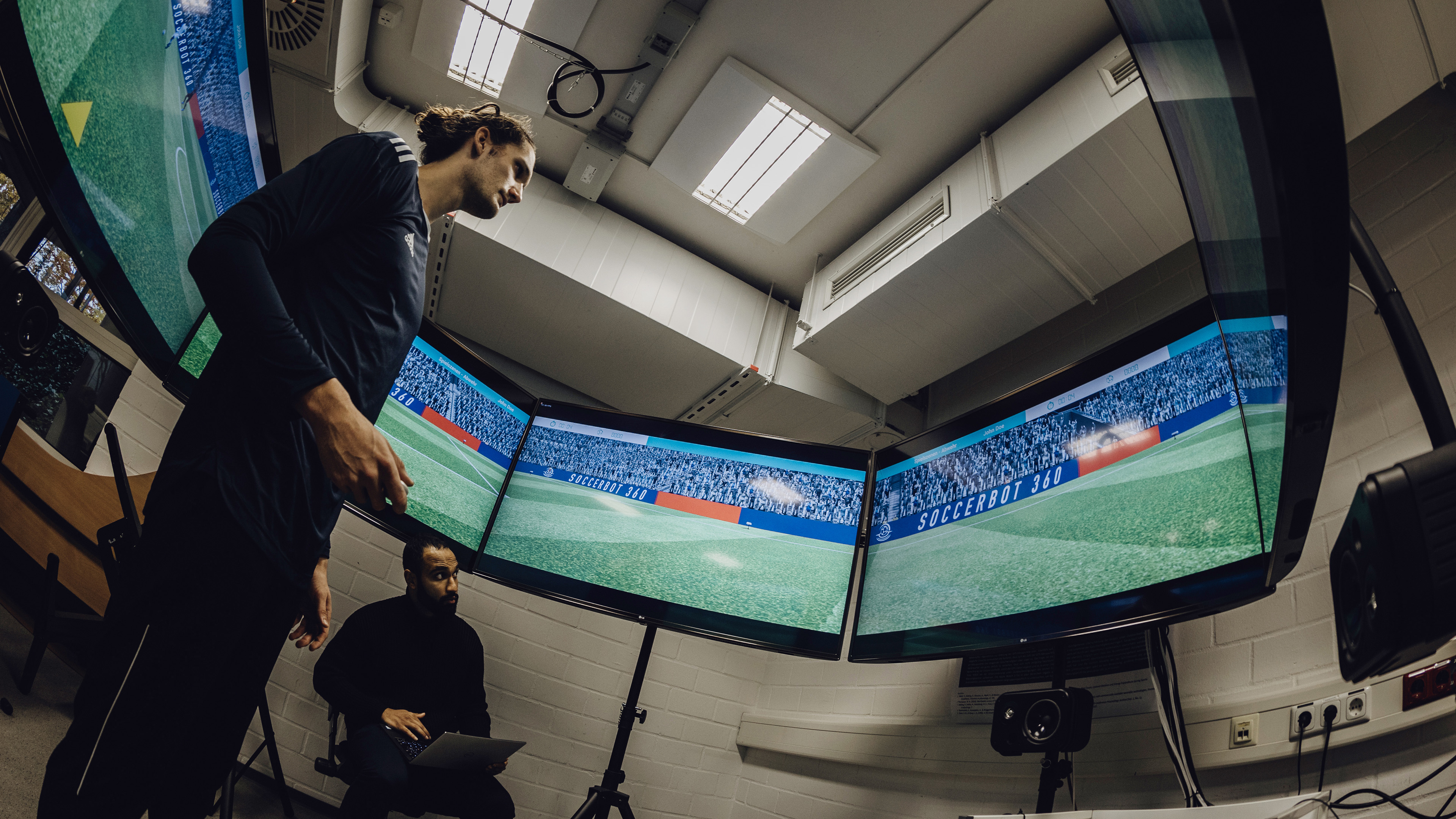 Foto von Mann der in der Mitte von sechs kreisförmig aufgestellten Bildschirmen mit Fußballsimulation steht
