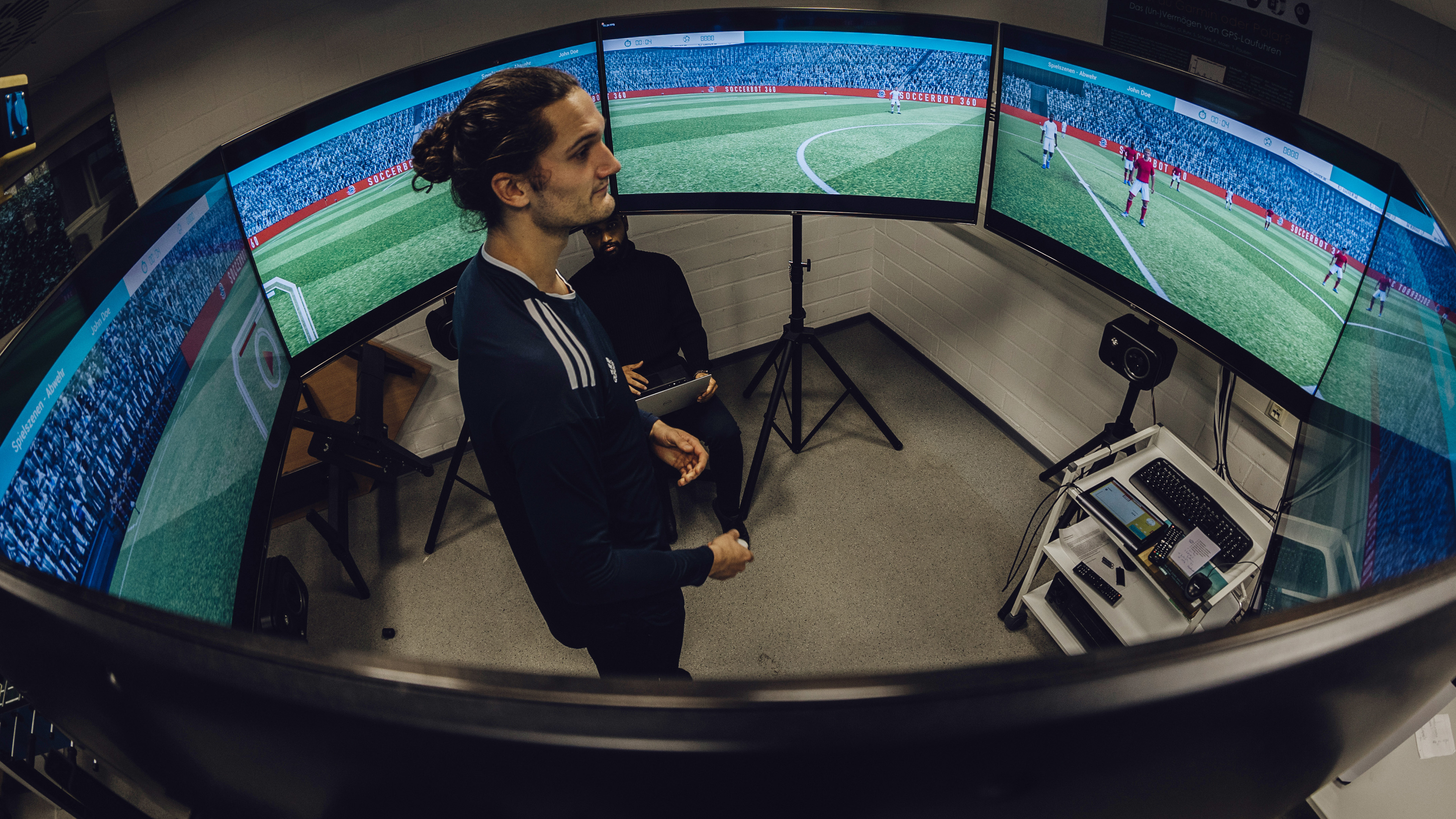 Foto von Mann der in der Mitte von sechs kreisförmig aufgestellten Monitoren mit laufender Fußballsimulation blickt
