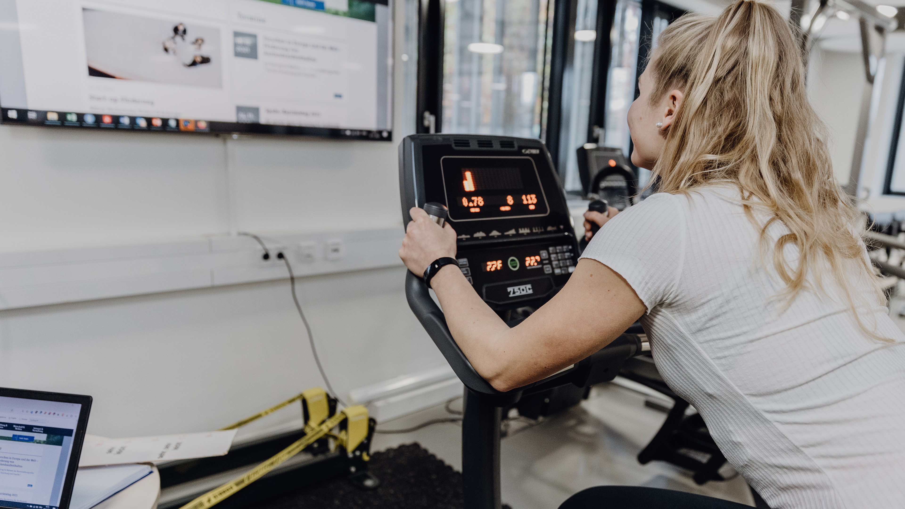 Foto von Frau die in Sportkleidung auf Ergometer sitzt und einen Bildschirm vor ihr betrachtet
