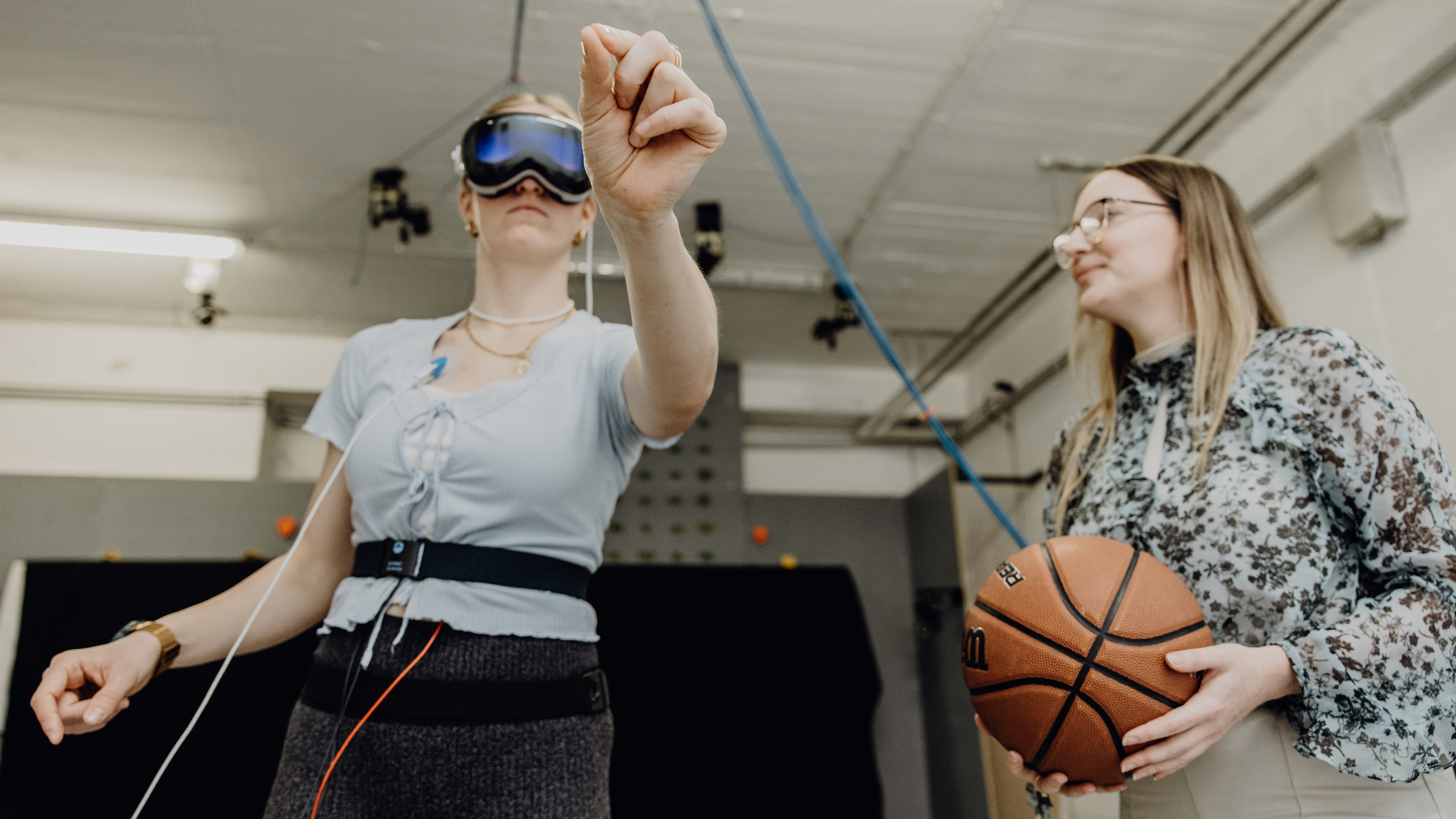 Probandin mit VR-Brille zeigt in leeren Raum, während forschende Person neben ihr einen Basketball hält
