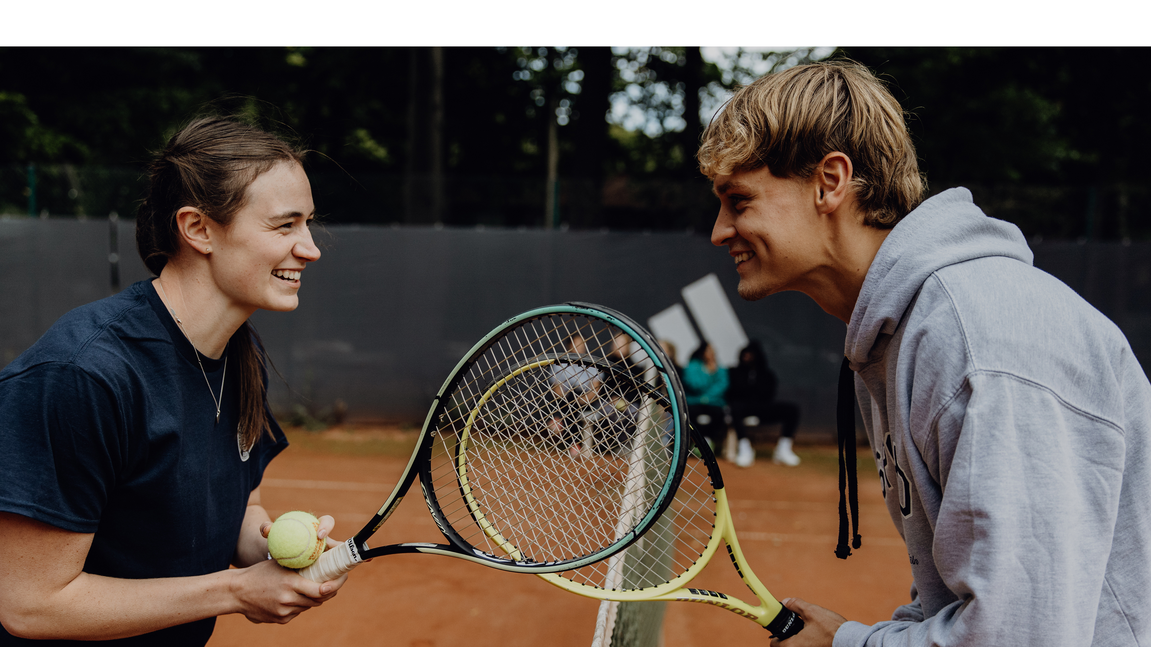 Foto von zwei Personen die sich lächelnd mit Tennisschlägern in der Hand gegenüber stehen
