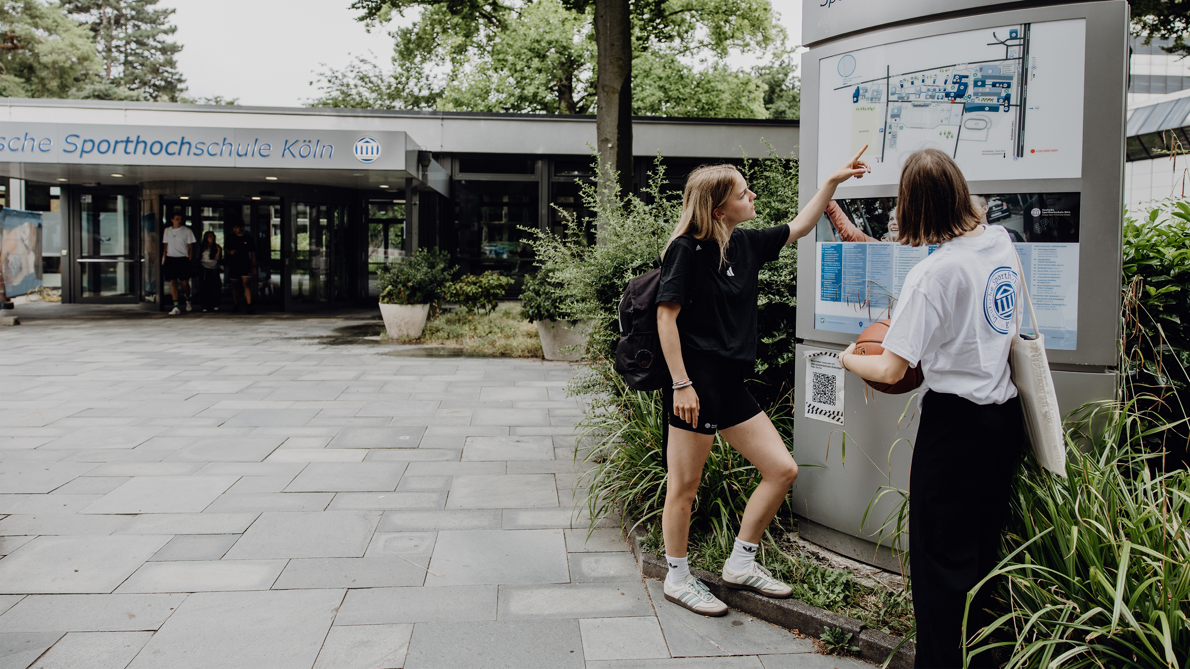 Zwei Studierende zeigen auf den Wegweiser im Eingangsbereich der Deutschen Sporthochschule
