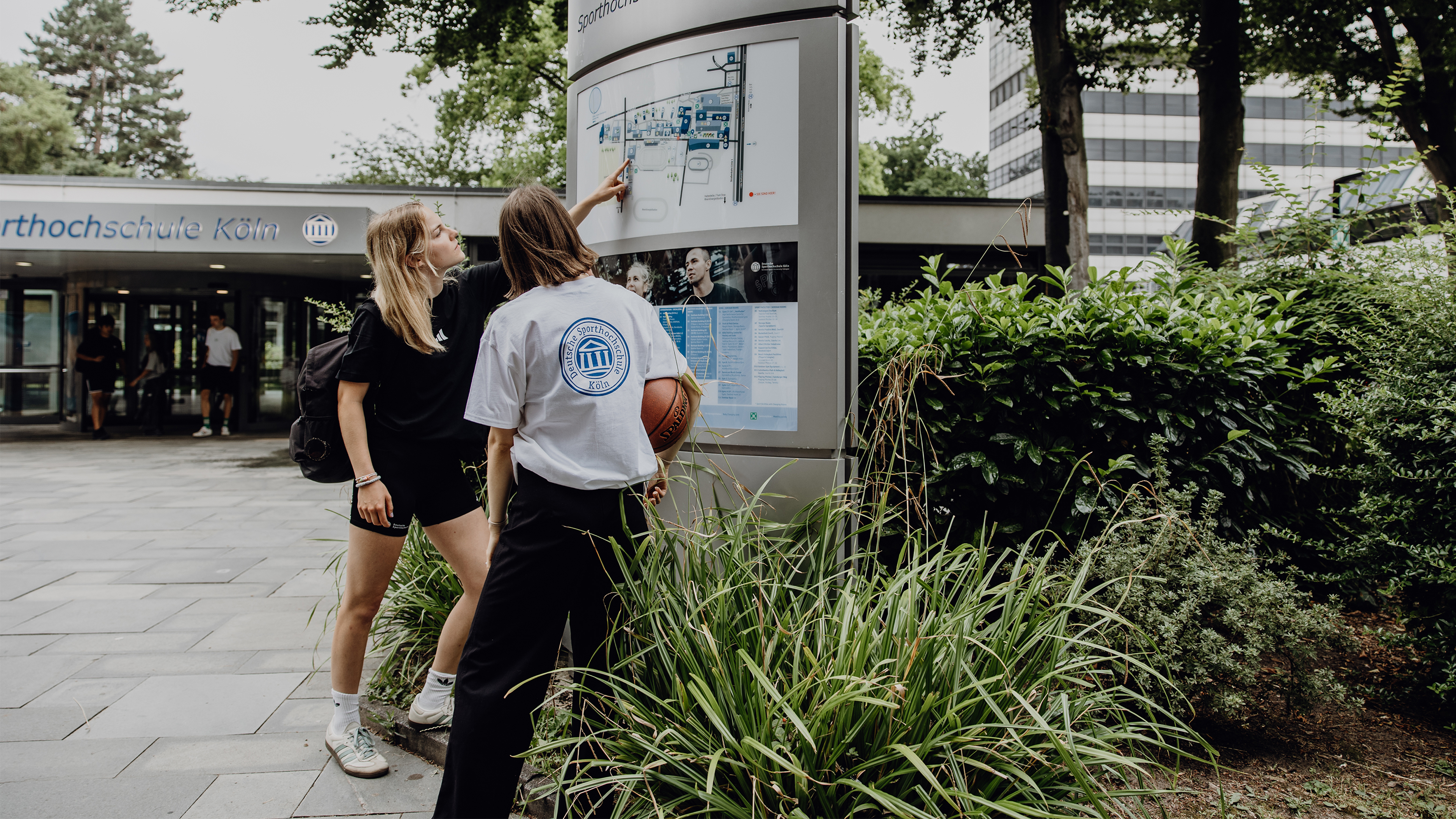 Zwei Studierende betrachten den Wegweiser im Eingangsbereich der Deutschen Sporthochschule
