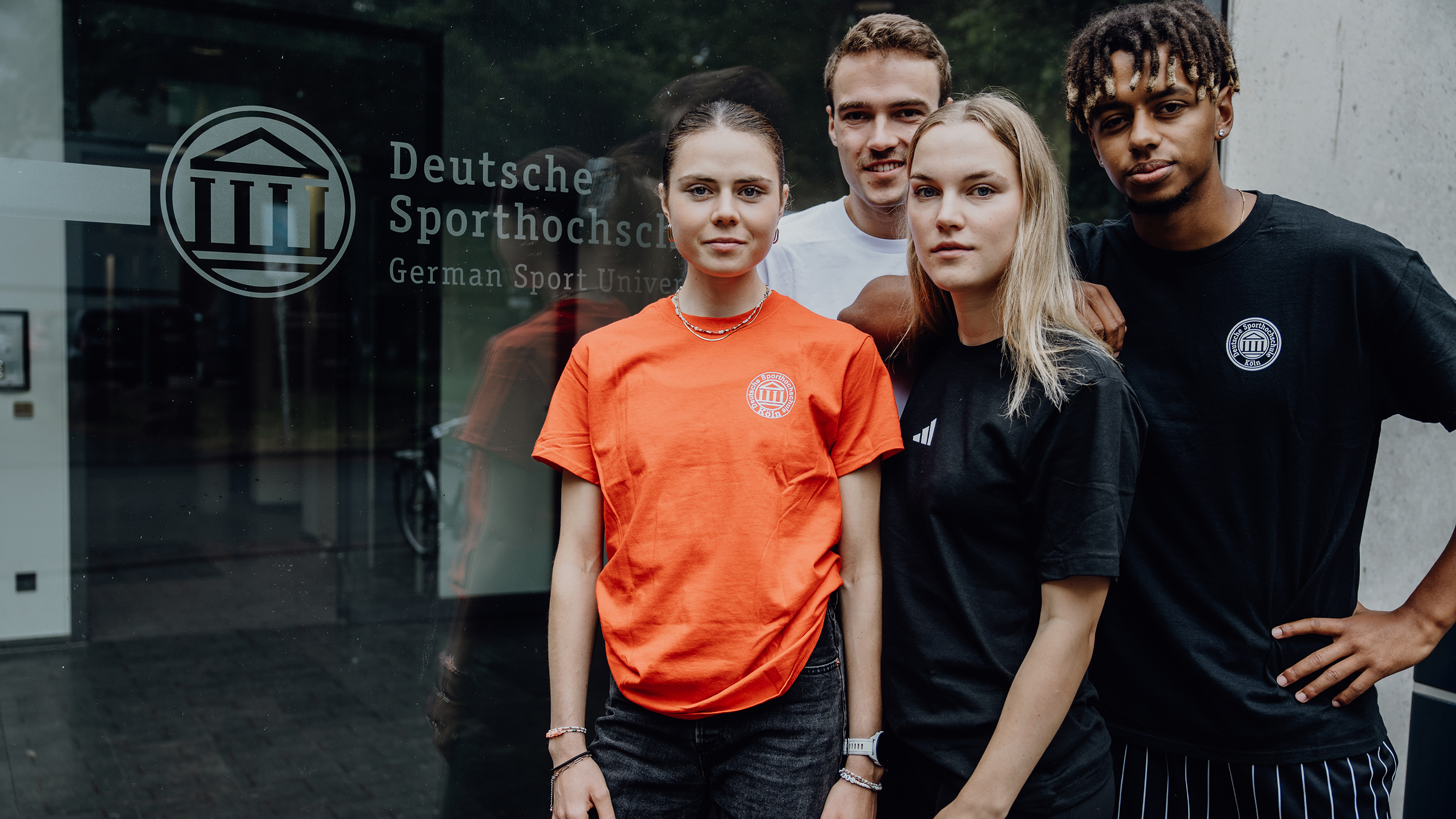 Vier Studierende stehen vor einer Glaswand mit der Aufschrift "Deutsche Sporthochschule"
