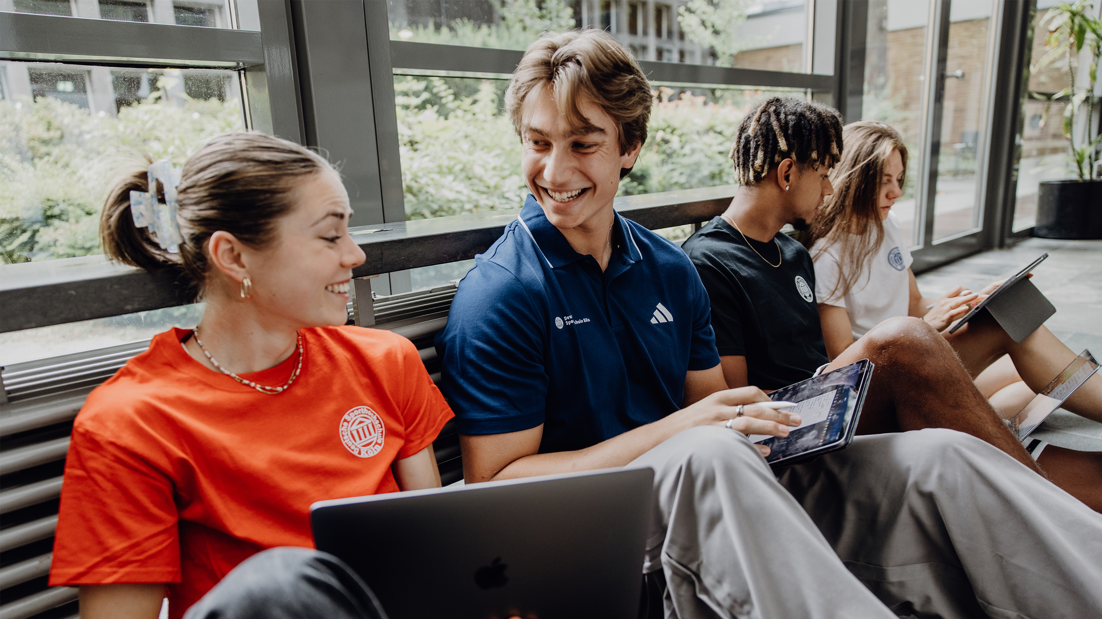 Vier Studierende sitzen auf dem Boden des Foyers und arbeiten mit Laptops und Tablets.
