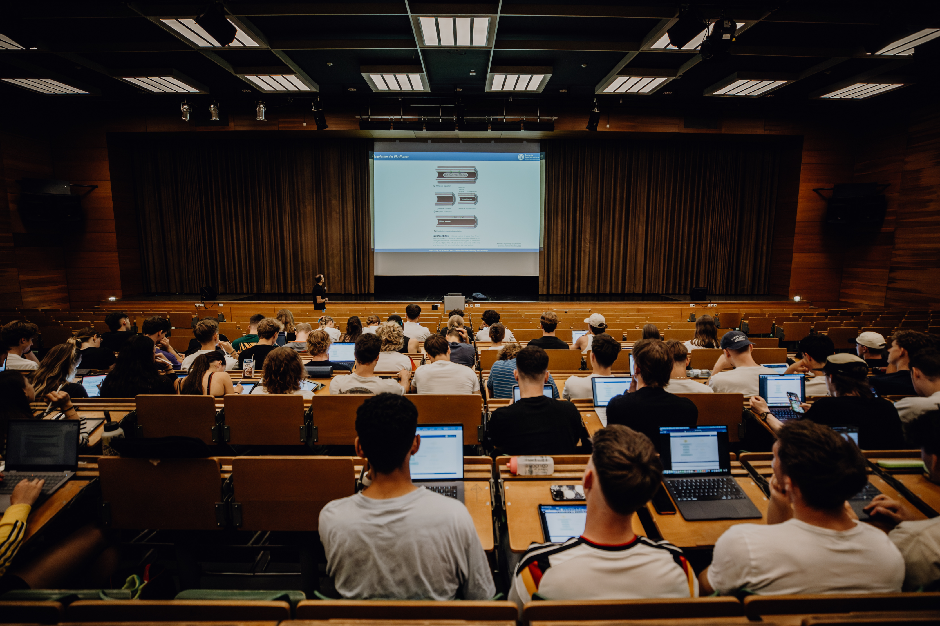 studierende sitzen im hörsaal und hören dem Dozenten zu. Viele haben Laptops vor sich.
